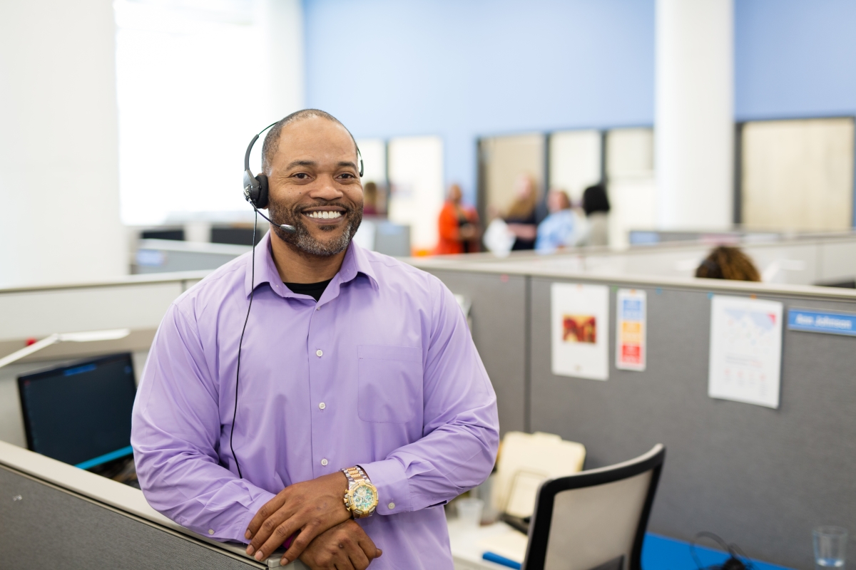 a smiling man wearing a phone headset stands in a cubicle